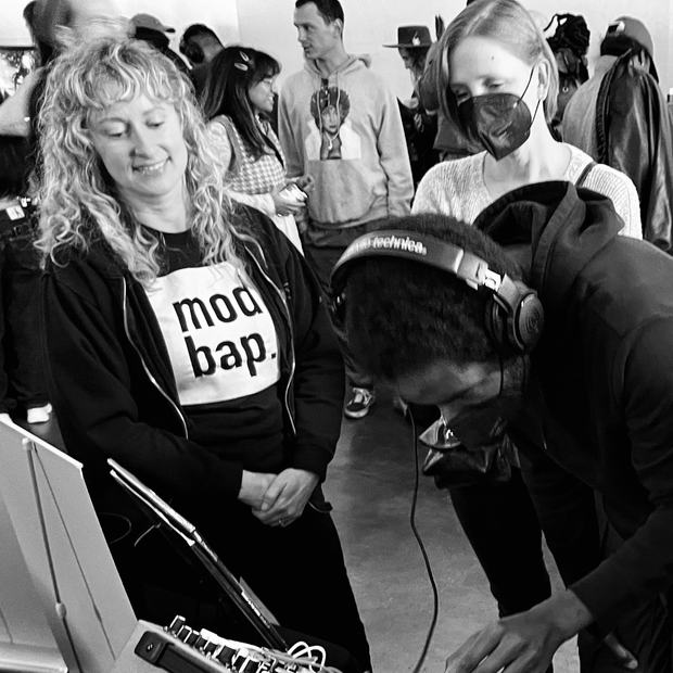 A visitor in a Modbap tee beside someone auditioning a rack on headphones